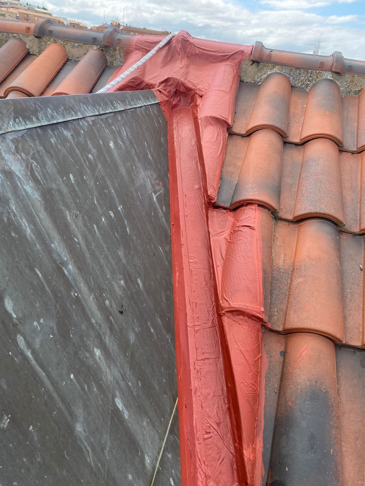 Canalón del tejado con membrana impermeabilizante roja entre tejas de terracota y un panel de remate de metal oscuro.
