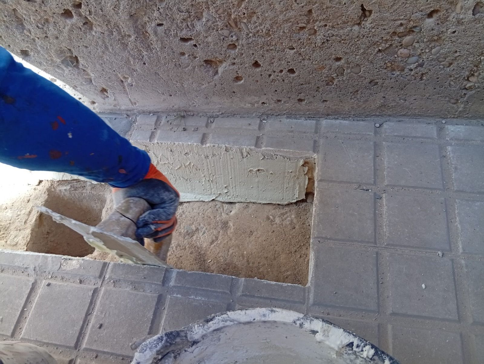 Una mano usa una paleta de mango azul para raspar el concreto de un agujero en un piso de baldosas rotas.