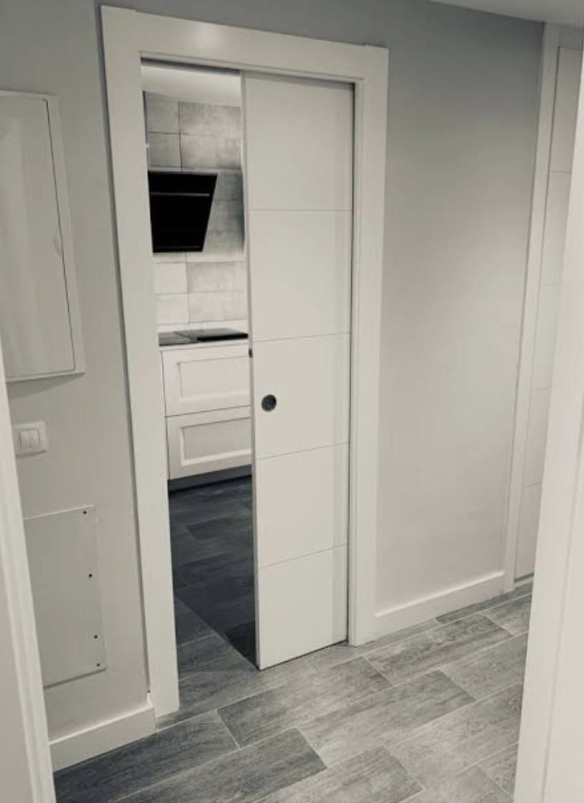 Pasillo blanco con puerta corredera abierta que da a una cocina luminosa con suelo gris.