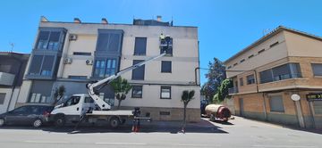 Una plataforma elevadora montada sobre camión está estacionada junto a un edificio blanco en una calle soleada