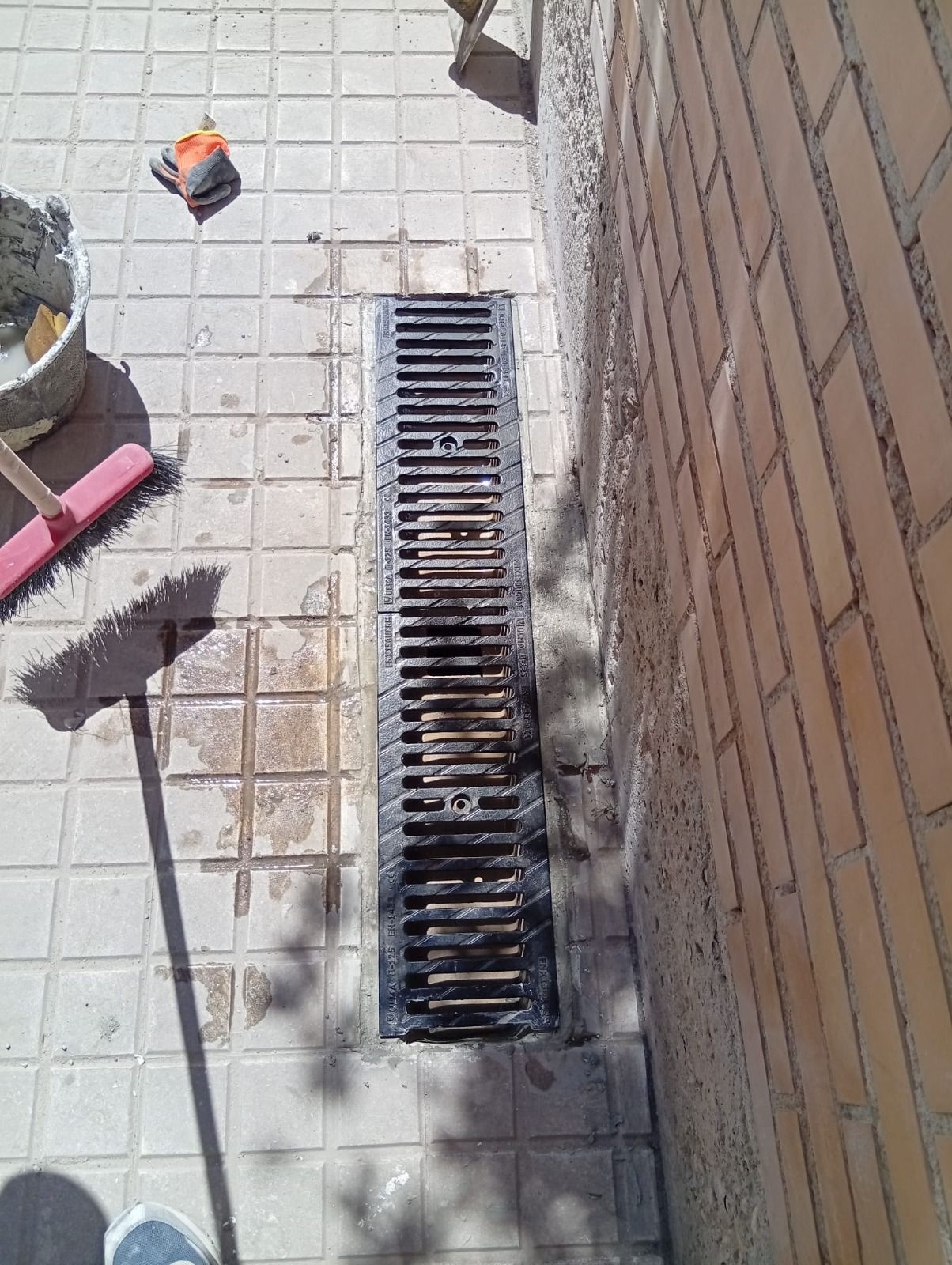 Estrecha rejilla de desagüe exterior instalada en un pavimento de baldosas junto a un muro de ladrillos, con escombros cerca.