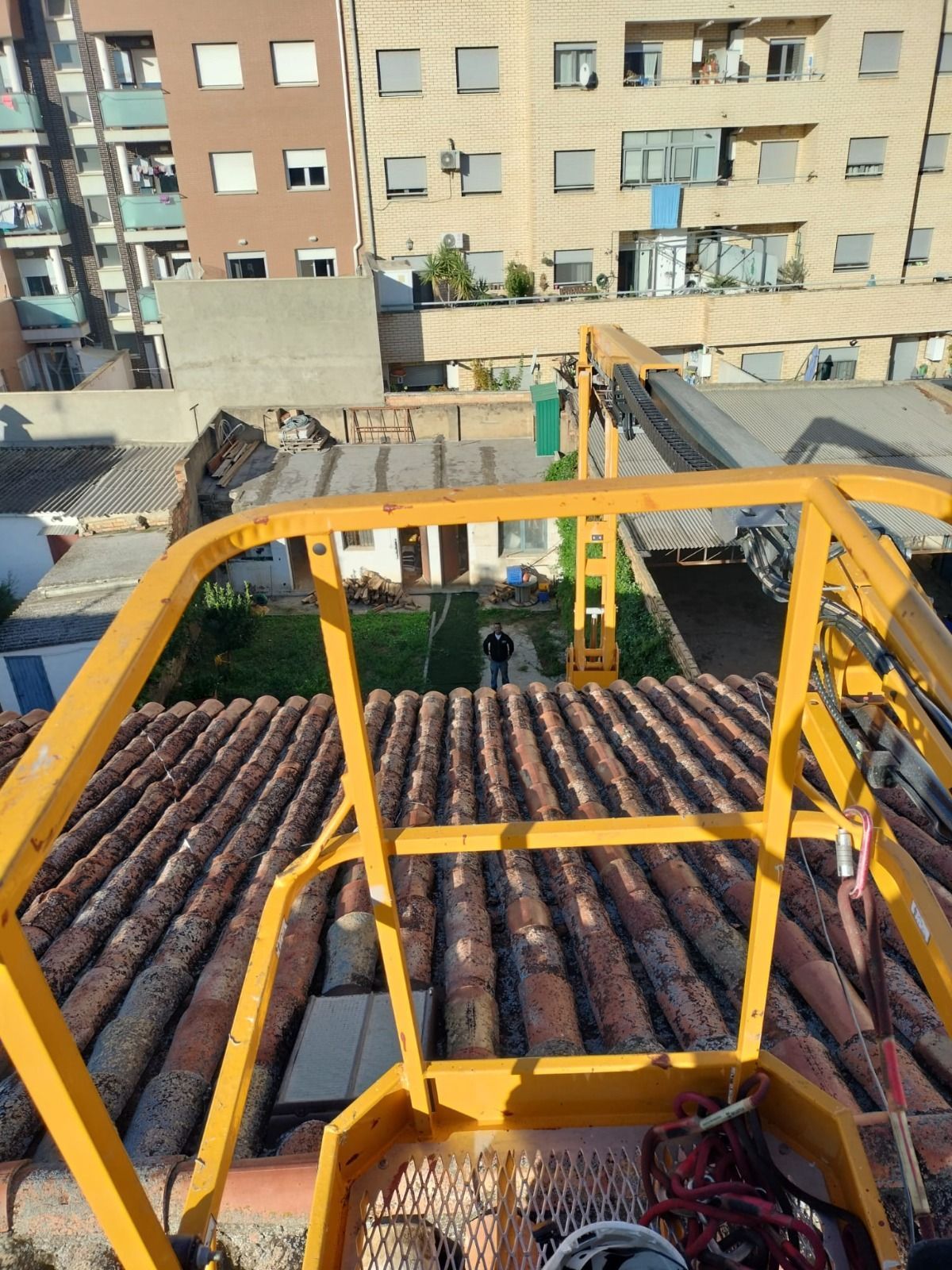 Vista desde la plataforma de una grúa amarilla sobre tejados de tejas y edificios de apartamentos en una zona urbana.