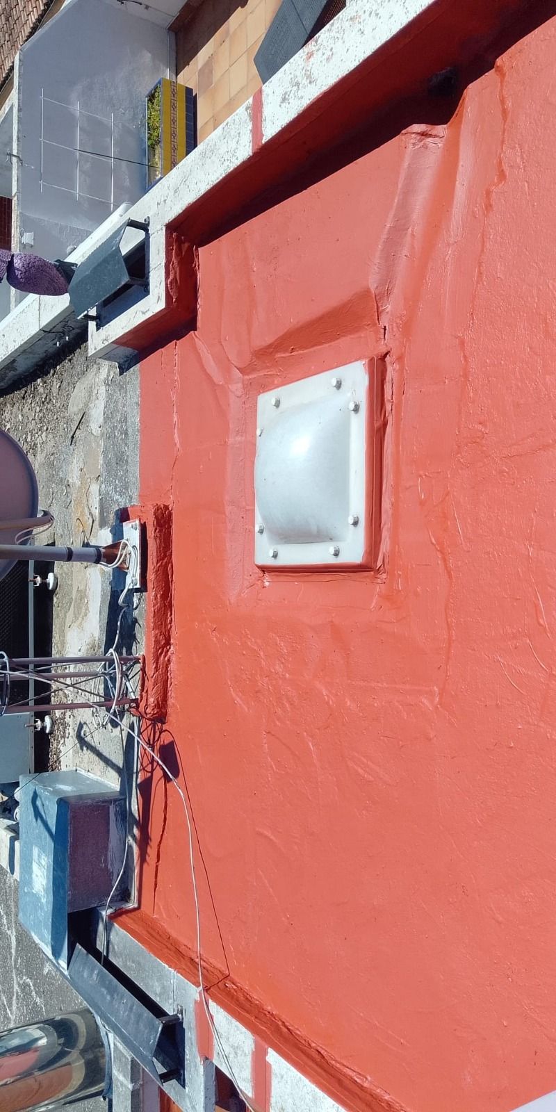 Fachada roja de un edificio con una rejilla de ventilación blanca y equipamiento en la azotea, junto a un callejón estrecho.