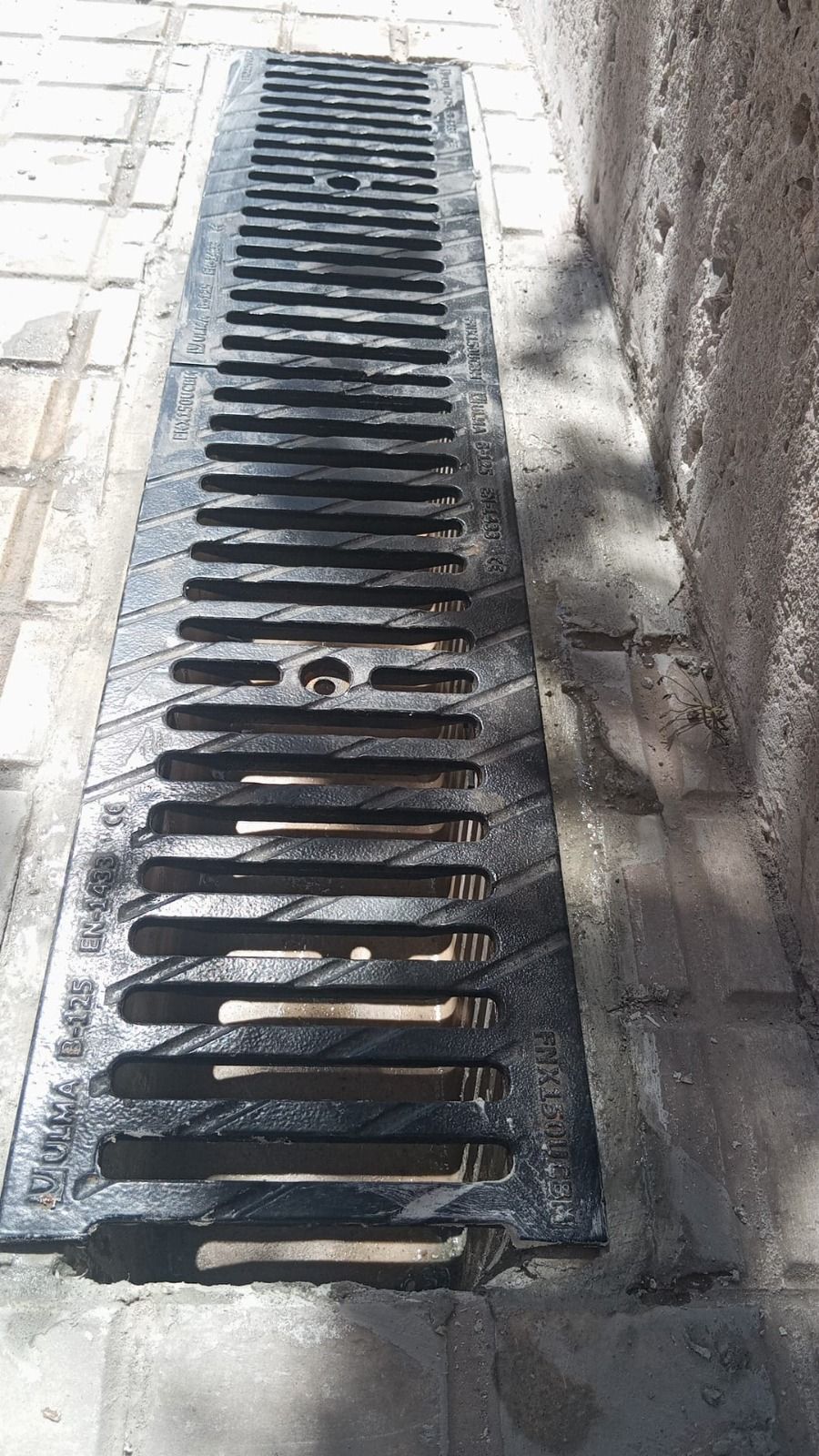 Rejilla metálica larga para desagüe pluvial colocada en una acera de ladrillo junto a un muro de hormigón.