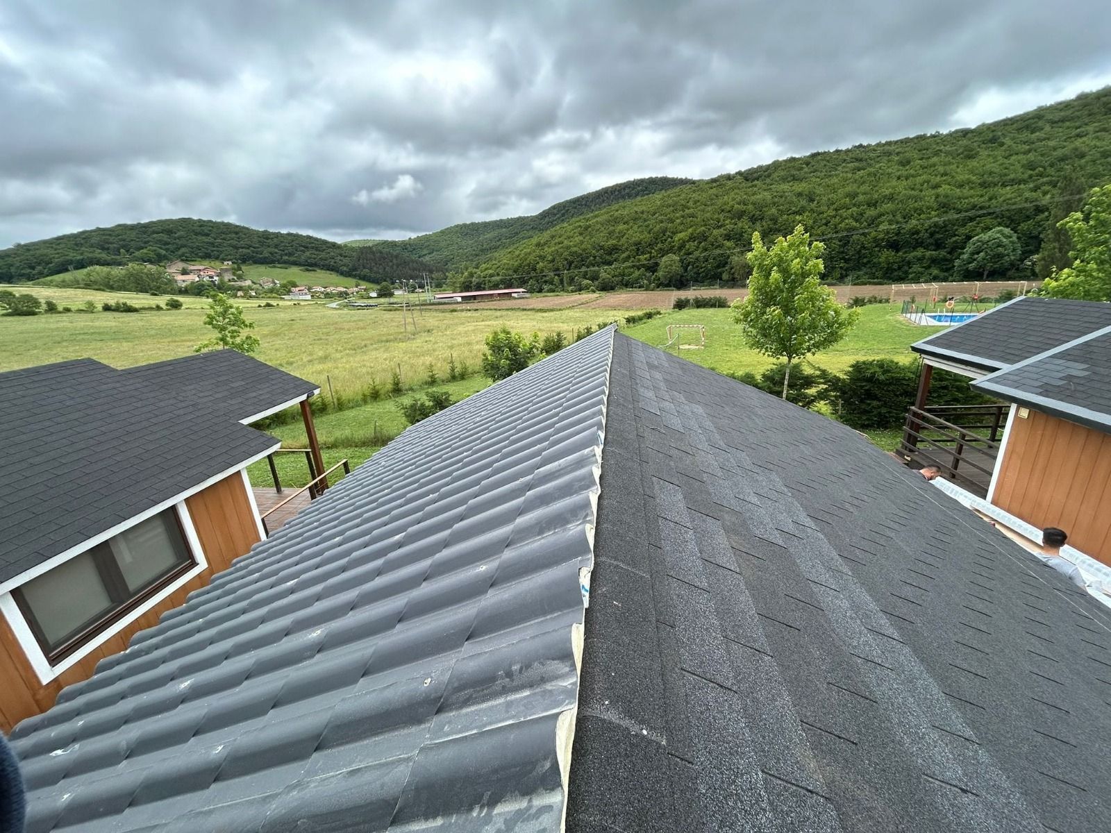 Vista desde la cumbrera de un tejado sobre casas grises, campos verdes y colinas boscosas bajo un cielo nublado.