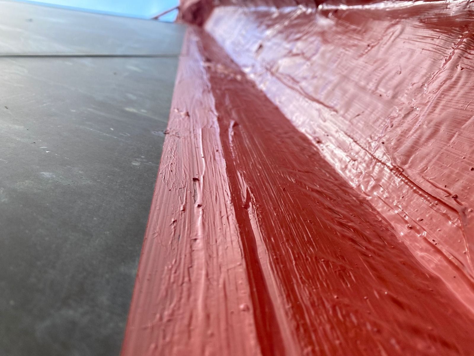 Primer plano de una superficie de madera pintada de rojo con pinceladas texturizadas junto a un suelo gris.