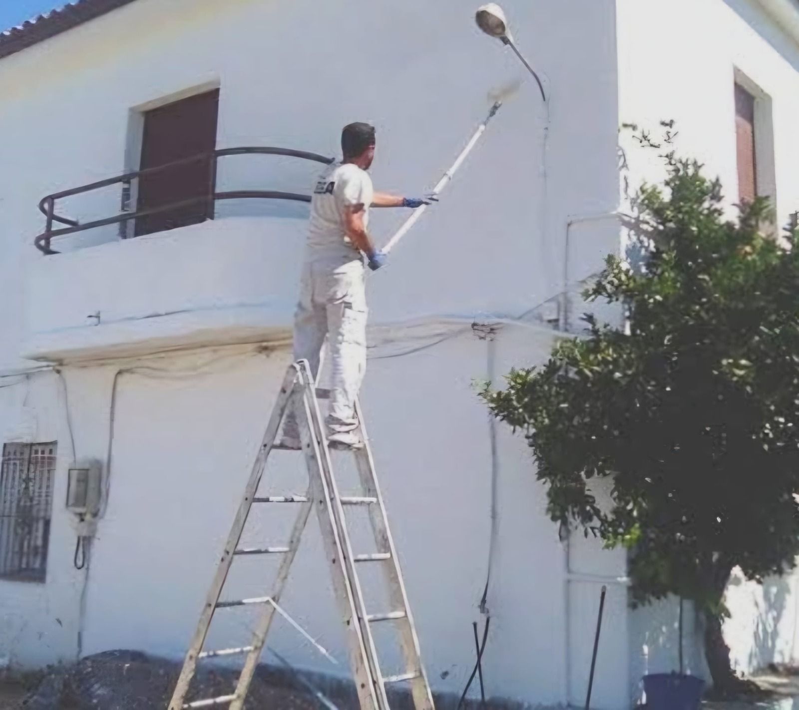 Una persona subida a una escalera pinta la fachada blanca de un edificio con un rodillo largo.