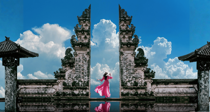 Person in pink standing between ornate stone temple gates reflected in water under a blue sky