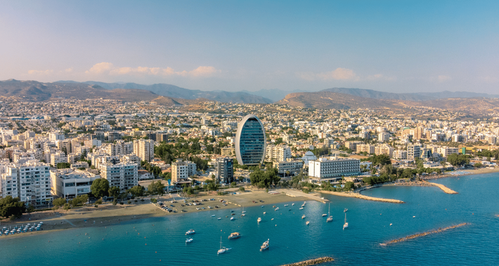 Aerial view of a sunny seaside city with a curved beach, turquoise water, and a prominent oval glass tower.