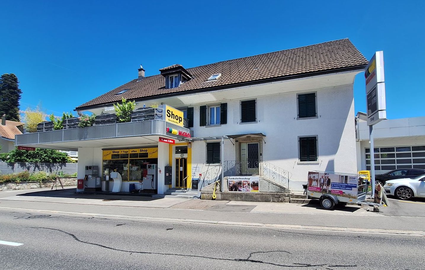 Weißes Gebäude mit Laden im Erdgeschoss, Balkon, dunkles Dach. In der Nähe geparkte Autos, sonniger Tag.