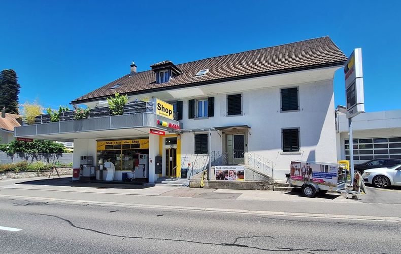 Weißes Gebäude mit Laden im Erdgeschoss, Balkon, dunkles Dach. In der Nähe geparkte Autos, sonniger Tag.