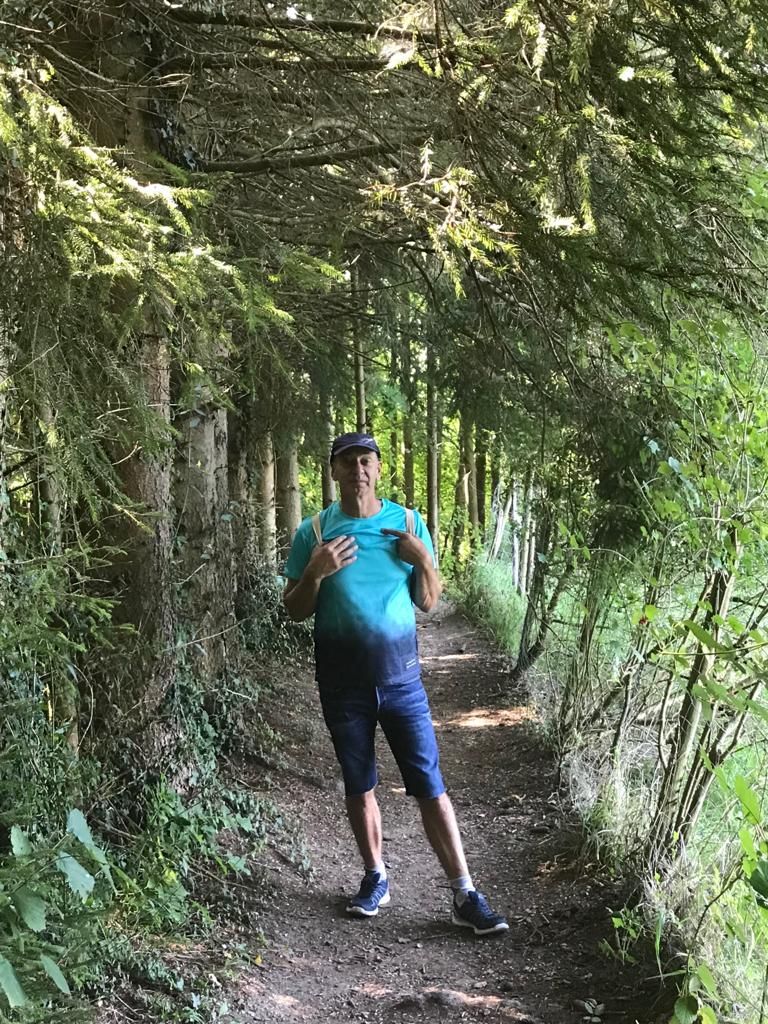 Ein Mann steht auf einem Feldweg, umgeben von Bäumen. Er trägt Blau und eine Mütze und deutet auf seine Brust.