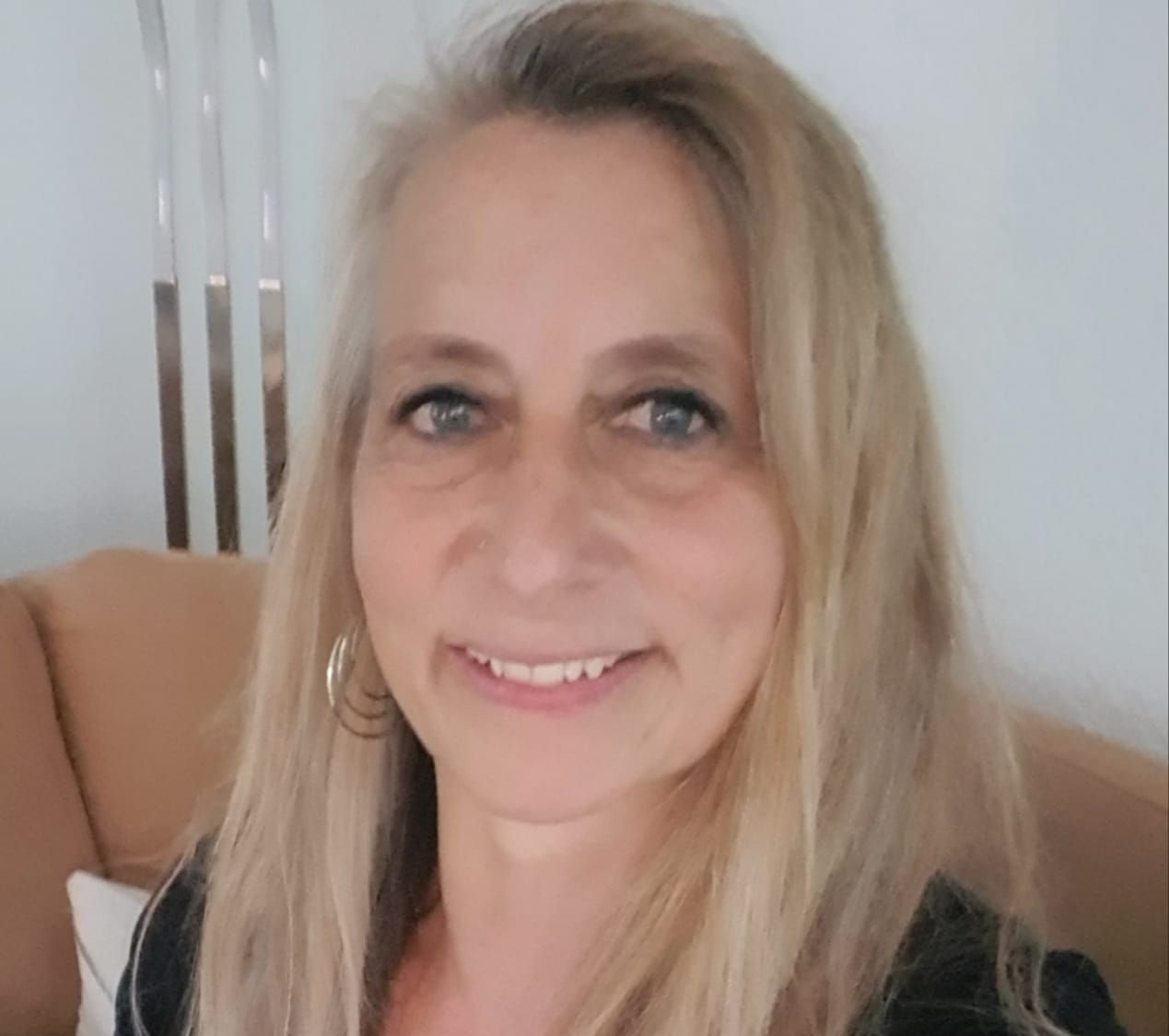 Frau mit langen blonden Haaren, lächelnd. Trägt Creolen und sitzt drinnen auf einem hellbraunen Sofa.