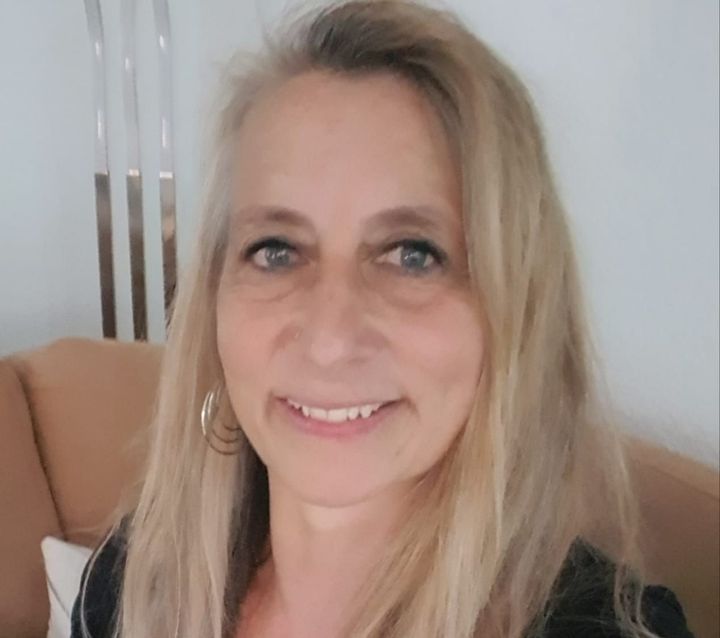 Frau mit langen blonden Haaren, lächelnd. Trägt Creolen und sitzt drinnen auf einem hellbraunen Sofa.