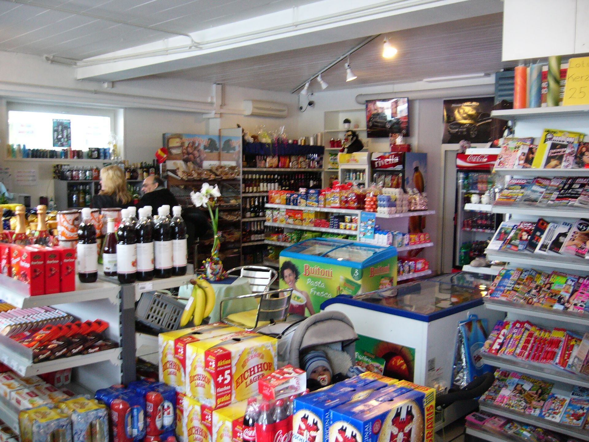 In einem kleinen Laden: Regale mit Getränken, Snacks, Zeitschriften und Eiscreme. Eine Person steht in der Nähe der Theke.