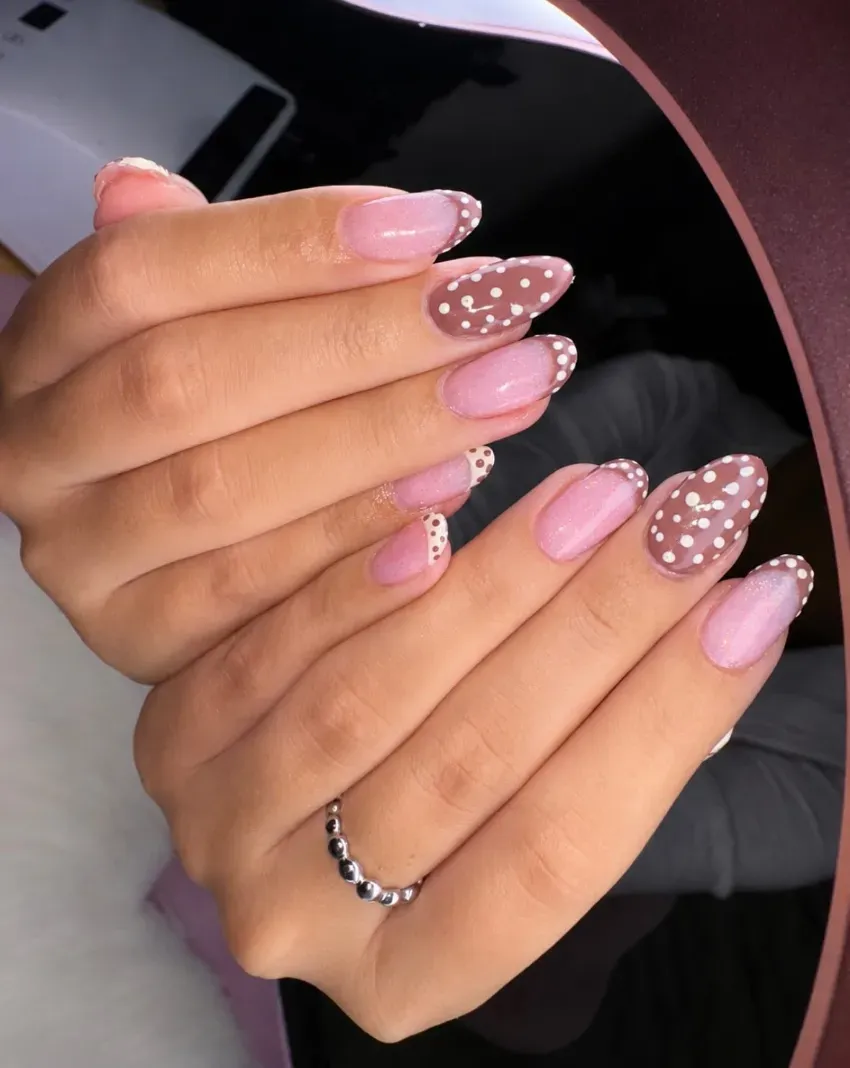 Manos con uñas pintadas con lunares rosas y marrones, mostradas sobre un espejo redondo.