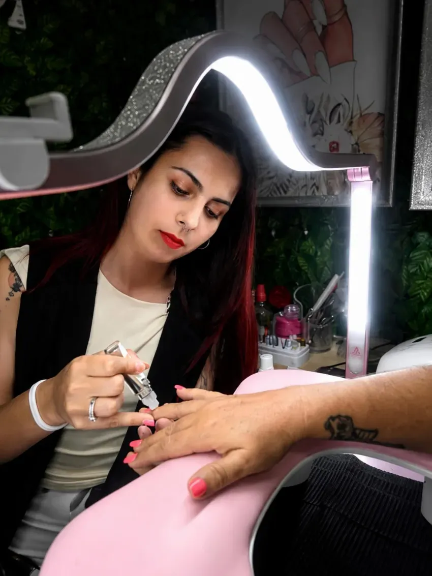 Una persona pinta las uñas de otra persona bajo una luz anular brillante, con un cojín rosa en primer plano.