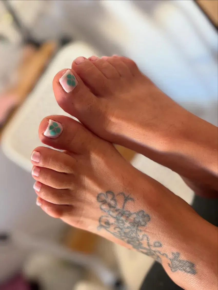 Dos pies descalzos con esmalte de uñas verde y un tatuaje floral en el tobillo sobre un fondo borroso.
