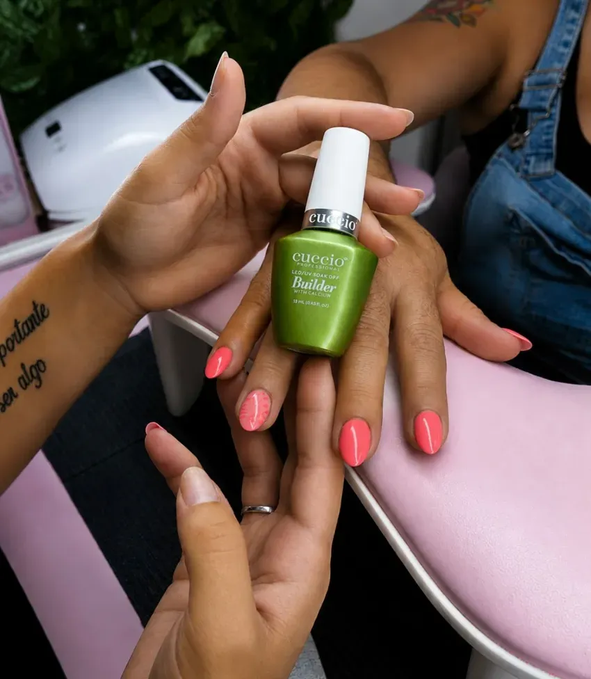 Una mano sostiene un frasco de esmalte de uñas verde sobre una estación de manicura rosa, dejando ver las uñas pintadas.