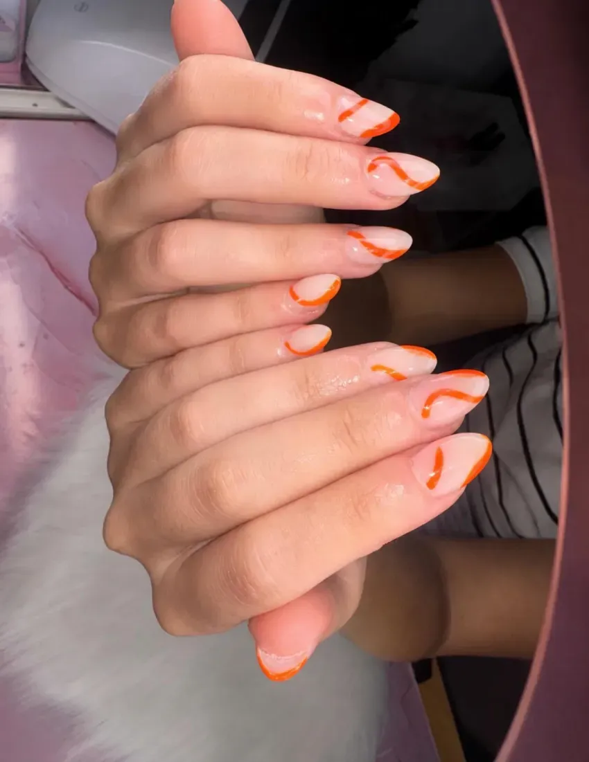 Primer plano de dos manos con uñas largas color nude y puntas de color naranja brillante, superpuestas en un entorno rosa suave.