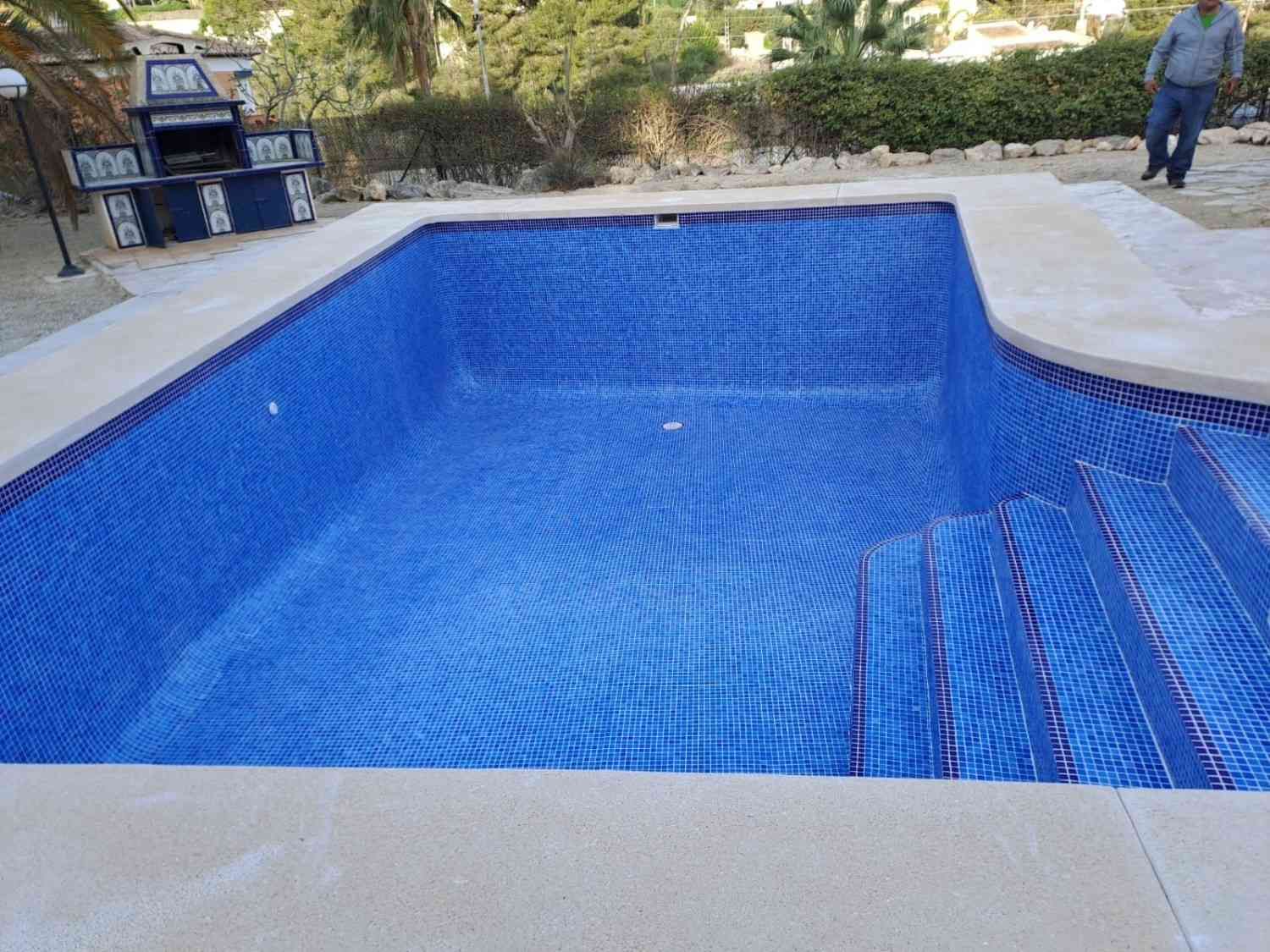 Piscina al aire libre con azulejos de mosaico azul, pasamanos y exuberante vegetación junto a un edificio.