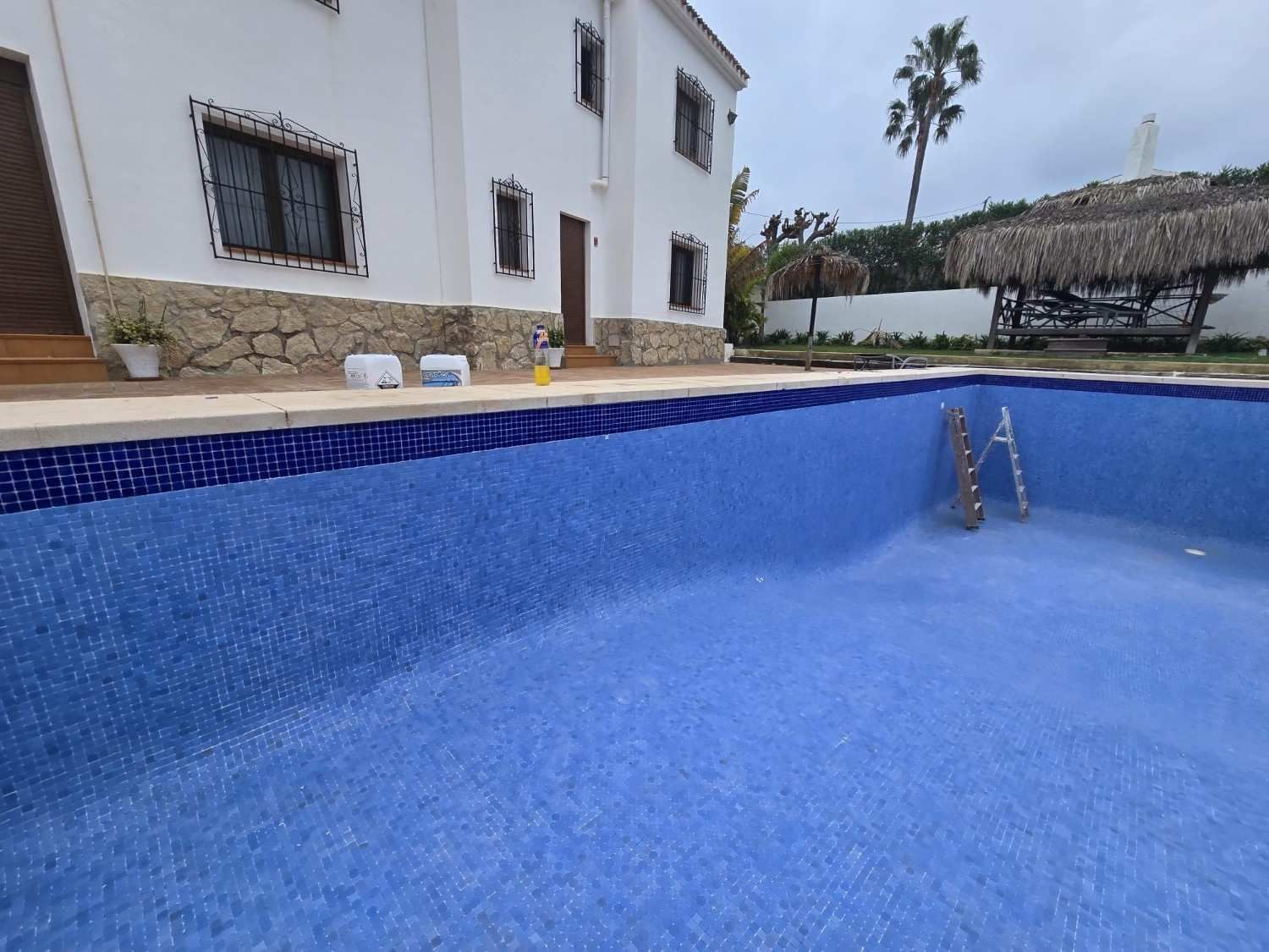 Piscina azul vacía junto a una casa blanca, con escalones y asientos de patio al fondo.