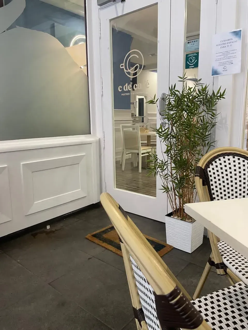 Entrada moderna de cafetería con puerta de cristal, sillas blancas, suelo a cuadros y una planta en maceta.