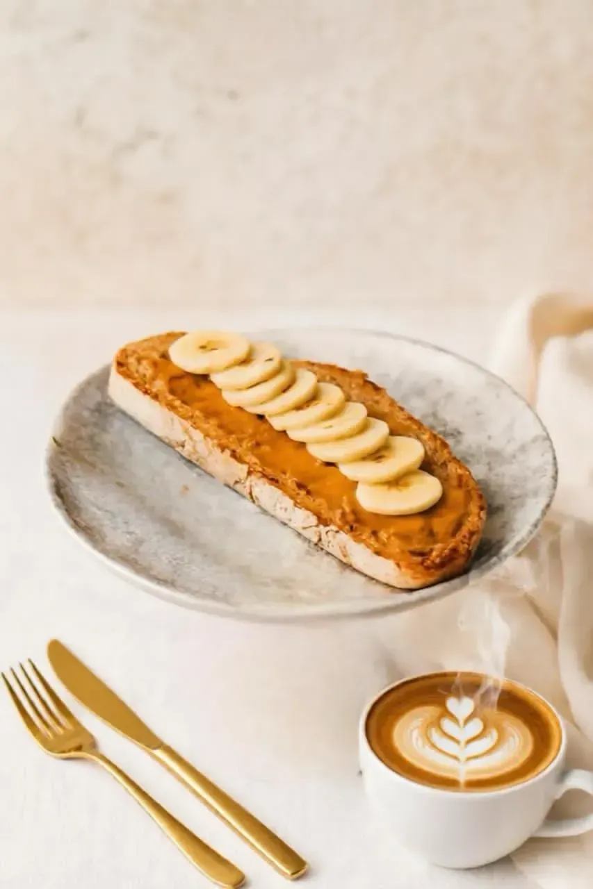 Pastel cubierto con rodajas de plátano en un plato, junto a un café con leche y cubiertos dorados sobre una mesa clara.