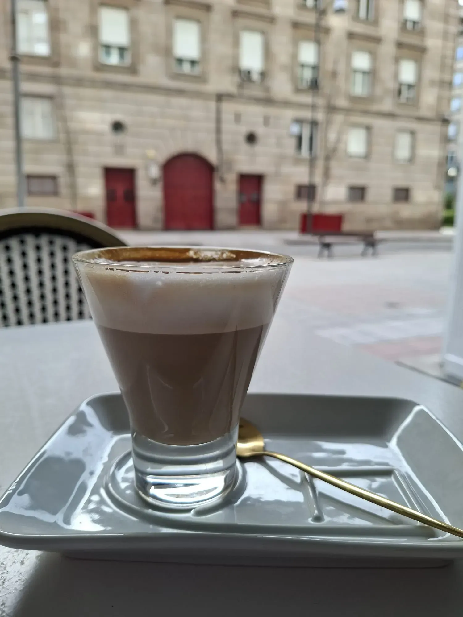 Vaso de café en capas sobre una bandeja con una cuchara, colocado al aire libre sobre una mesa de cafetería.