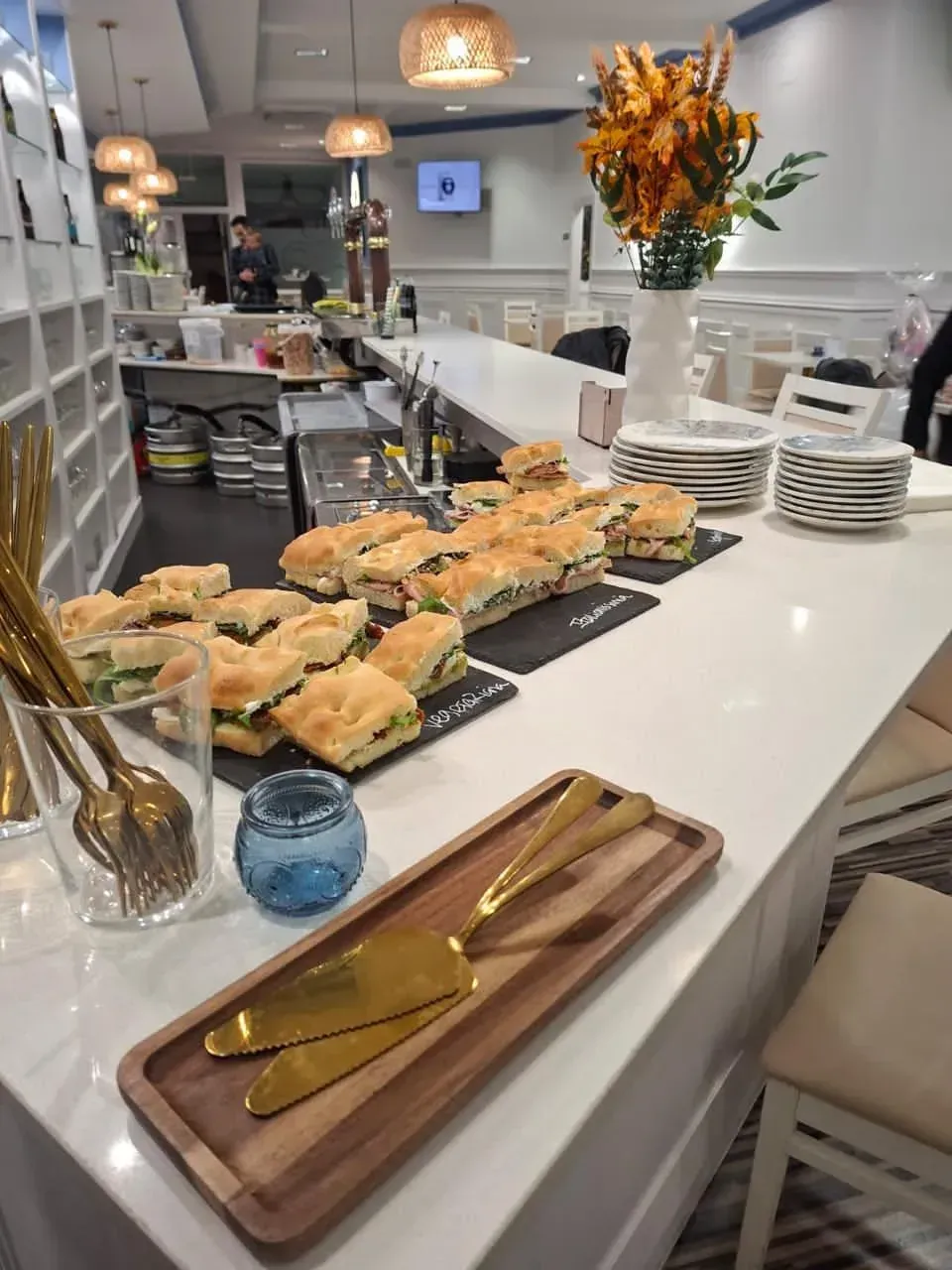 Mostrador de buffet con pasteles, platos y flores en un luminoso interior de cafetería.