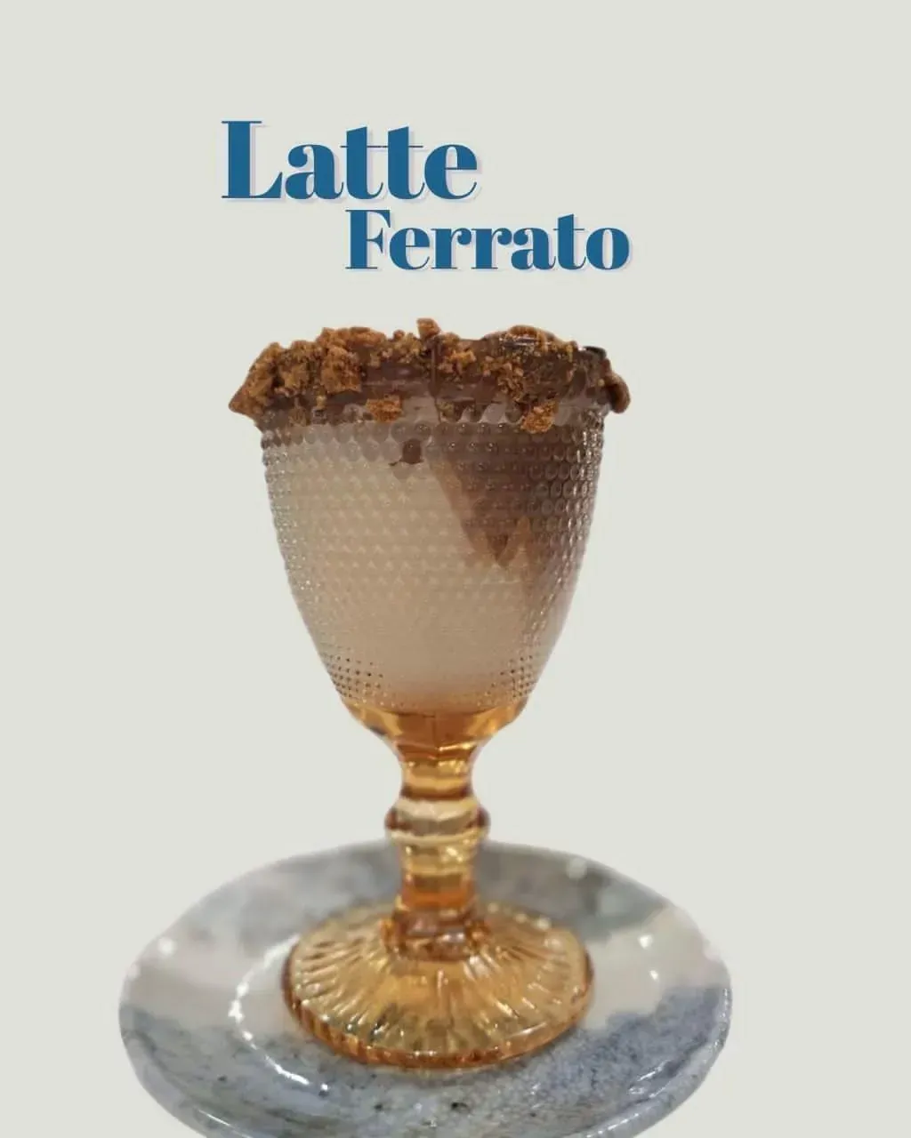 Latte Ferrato en una taza de cristal decorativa sobre un platillo, cubierto con migas de chocolate.