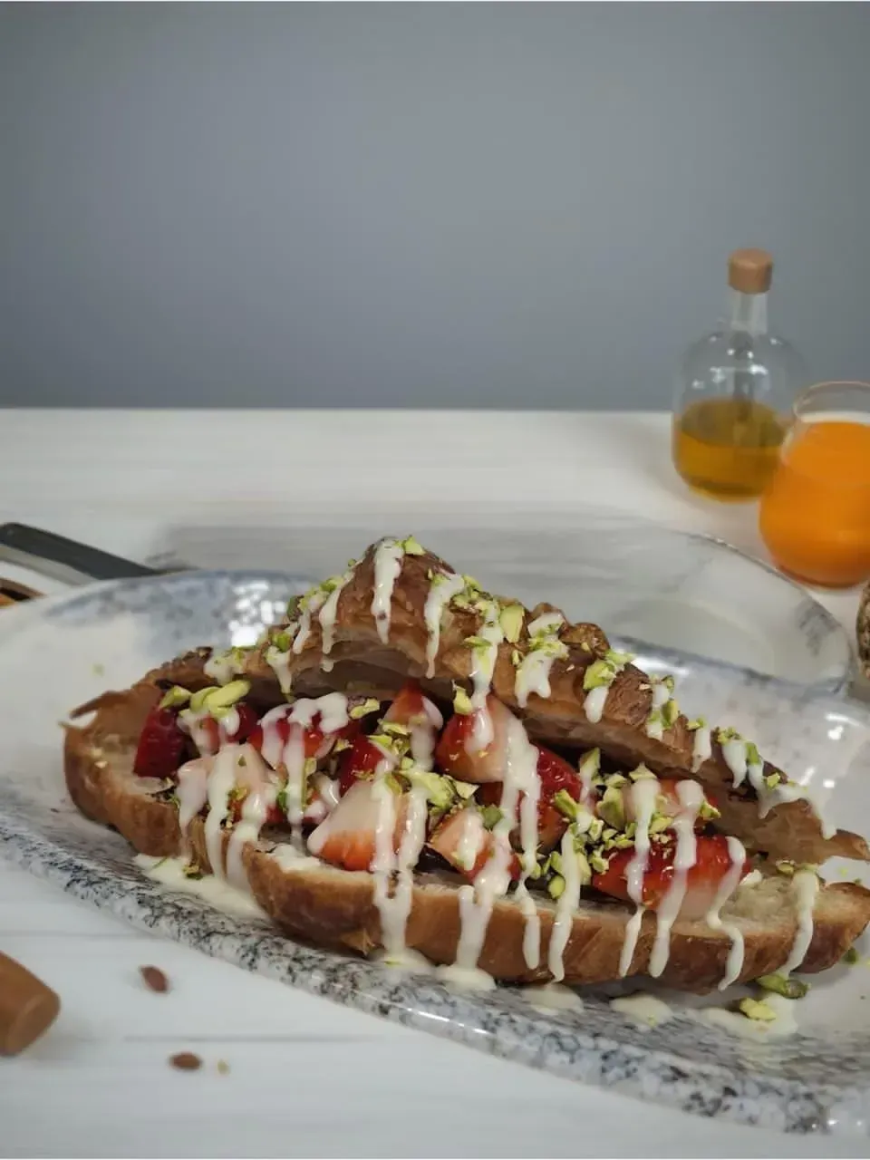 Un perrito caliente con salsas rociadas sobre una mesa, con una botella y un vaso al fondo.