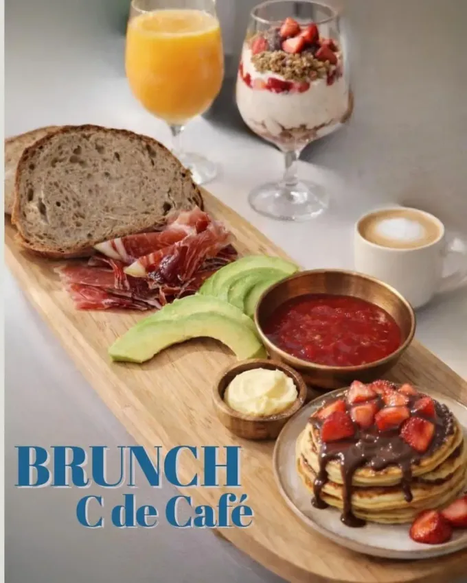 Desayuno buffet con tostadas de aguacate, sopa, tortitas, café, zumo de naranja y embutidos servidos en una tabla de madera.