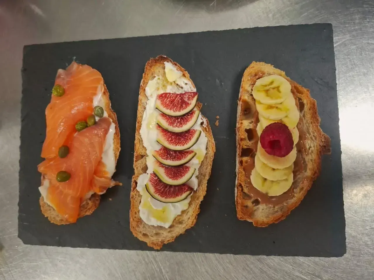 Tres tostadas abiertas: salmón, higos con crema y plátano con frutos rojos sobre una pizarra negra.