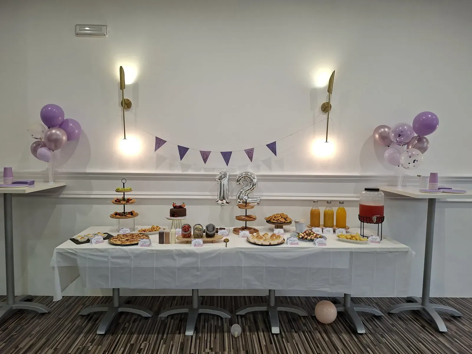 Mesa de postres decorada con globos morados, guirnaldas y pasteles variados en una habitación luminosa.