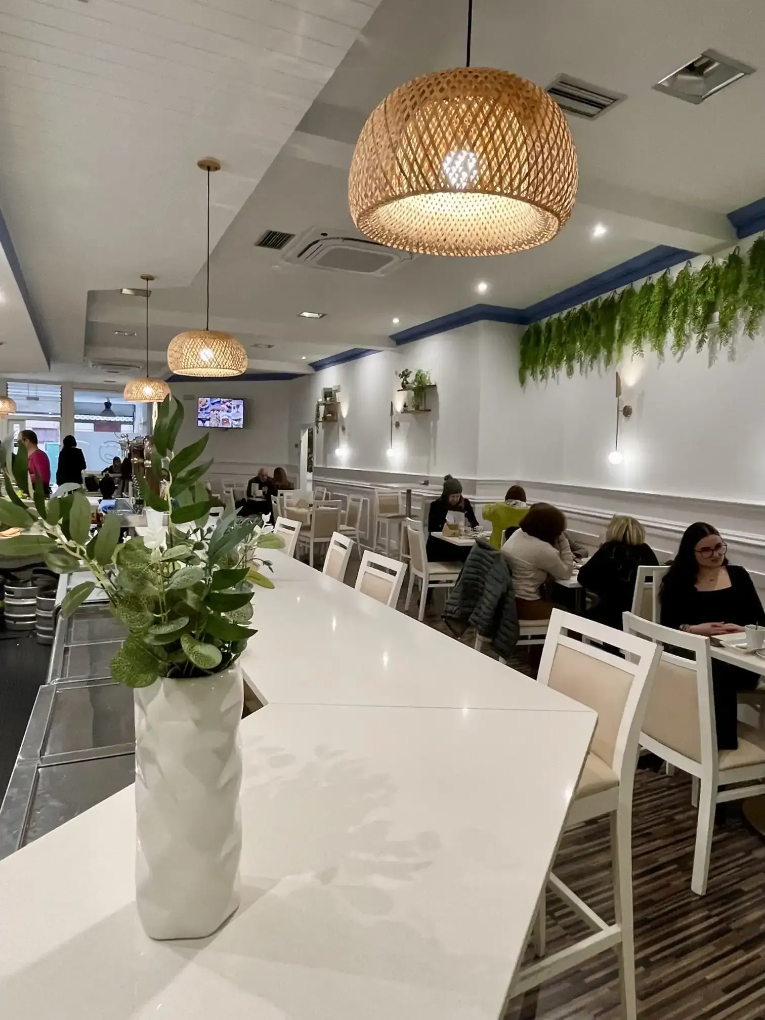 Interior luminoso de una cafetería con lámparas colgantes de mimbre, mesas blancas y gente sentada a lo largo de la pared.