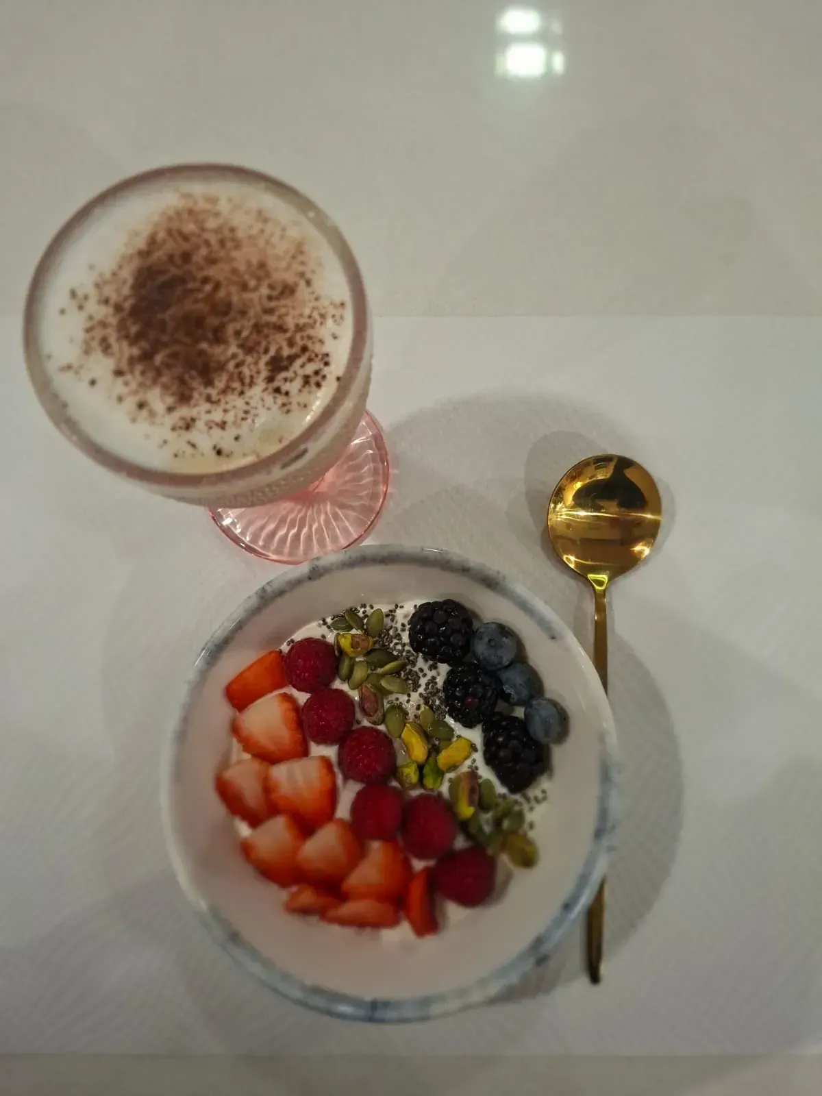 Un vaso de bebida espumosa y un tazón de yogur con frutas sobre una mesa blanca con una cuchara dorada.