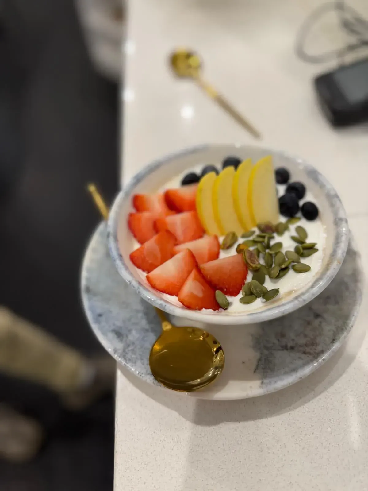 Bol de fruta y yogur cubierto con fresas, rodajas de plátano, arándanos y semillas en un plato con miel.