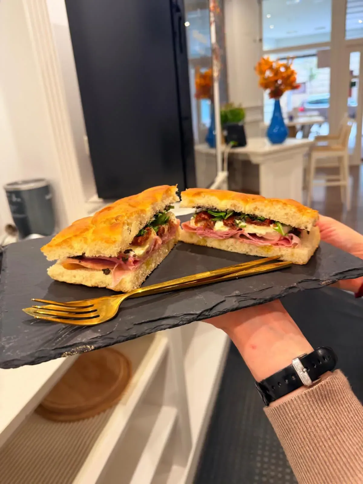 Una mano sostiene un plato de pizarra con un sándwich cortado, una encimera de cocina blanca y un jarrón al fondo.