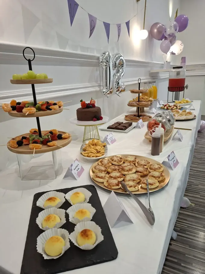 Buffet de postres con cupcakes, tartas, pasteles y bandejas de frutas en soportes y bandejas blancas de varios niveles en un salón de fiestas.