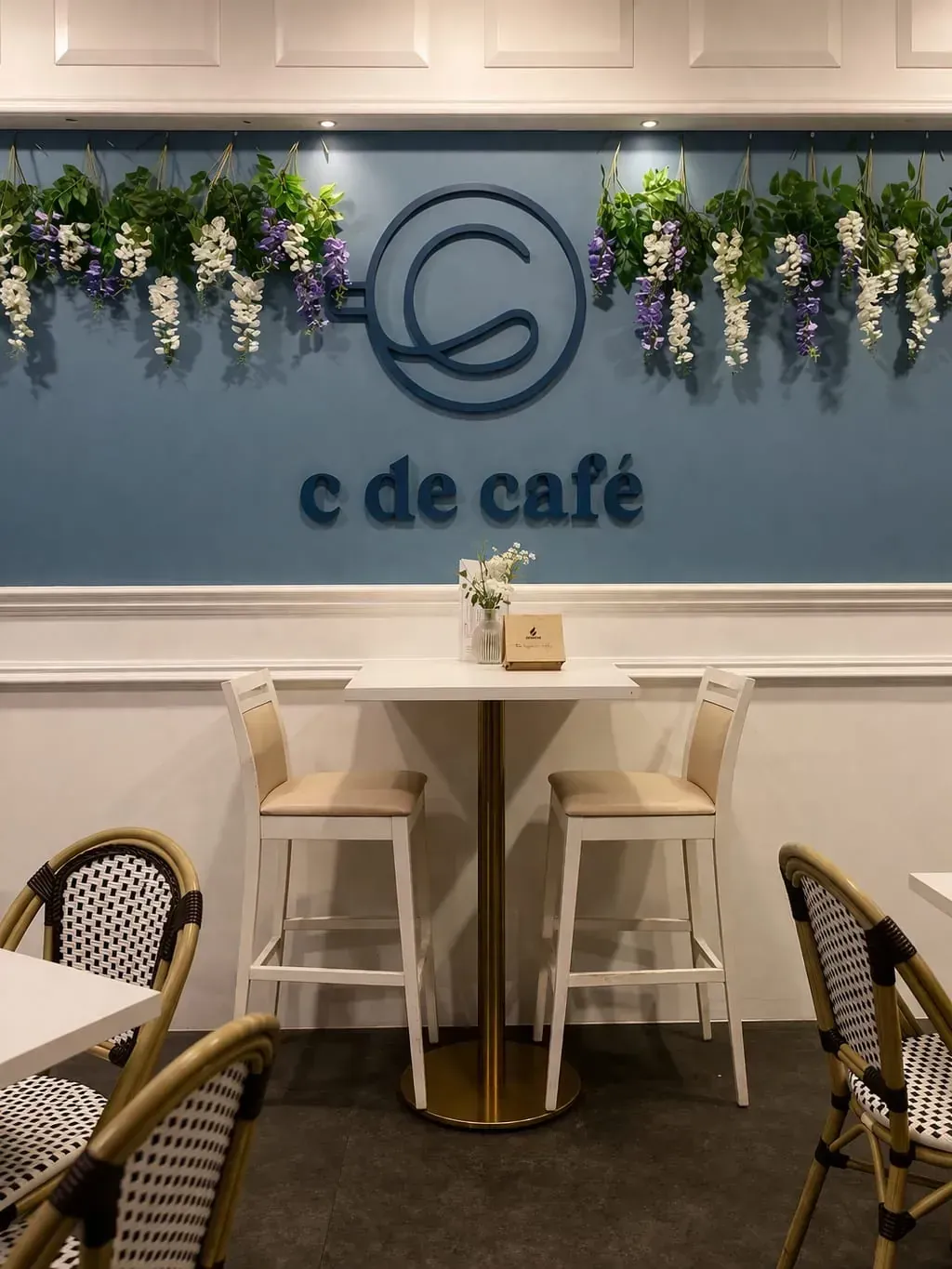 Interior de cafetería con logotipo azul en la pared, flores colgantes y una pequeña mesa redonda con taburetes de bar.