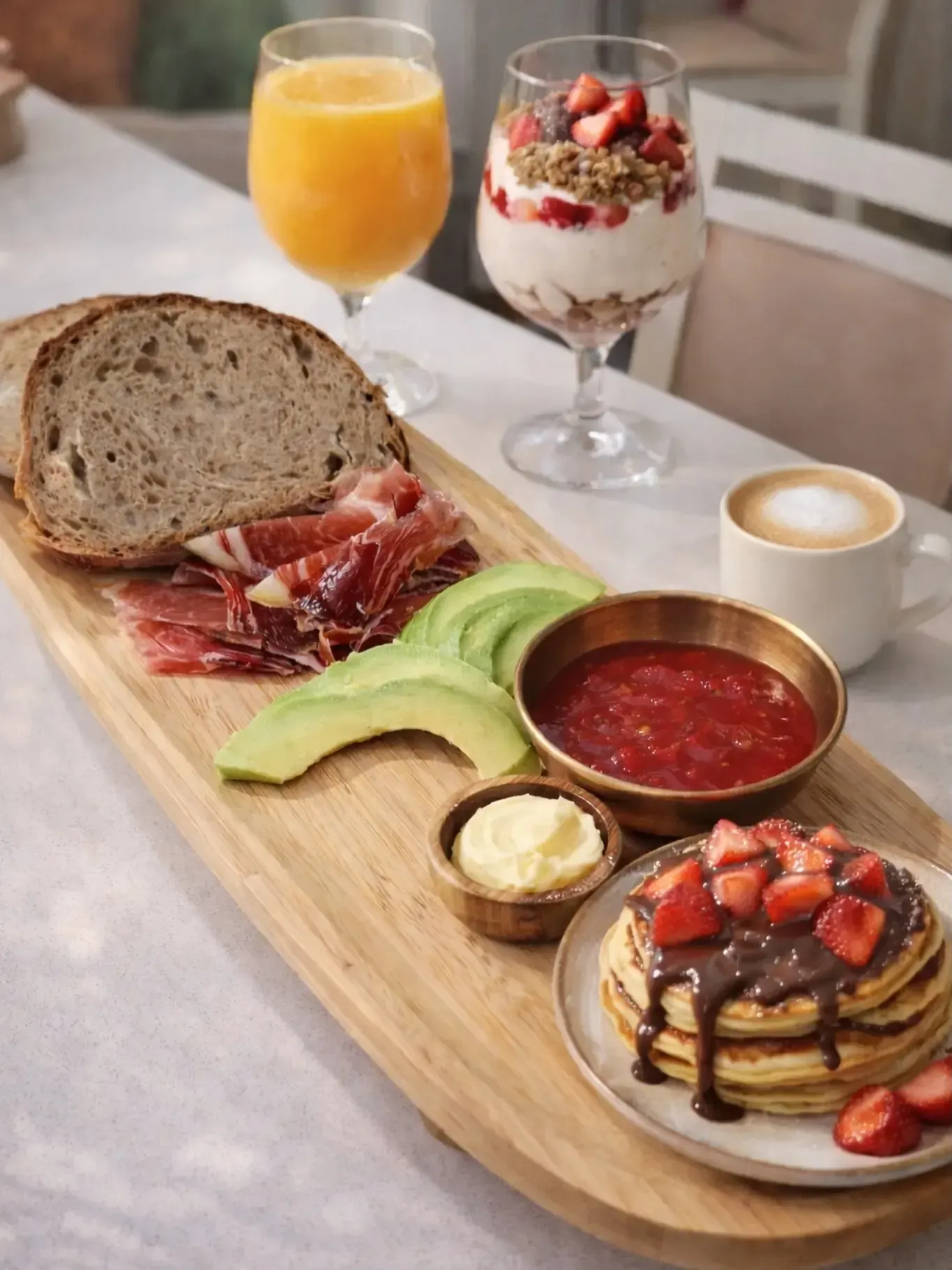 Desayuno completo con pan, jamón serrano, aguacate, frutos rojos, tortitas, zumo y café sobre una tabla de madera.