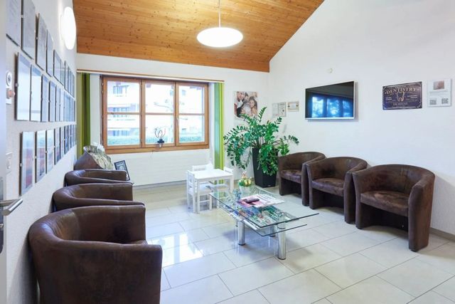 Wartezimmer mit braunen Sesseln, weißem Tisch, Fernseher und gerahmten Fotos.