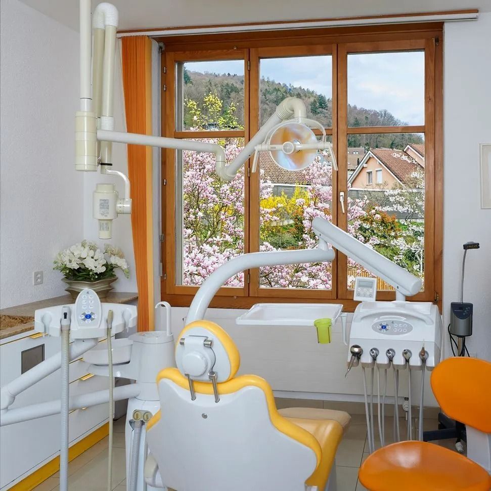 Zahnarztpraxis mit gelb-weißem Stuhl, Ausstattung und Fensterblick auf Häuser und Bäume.