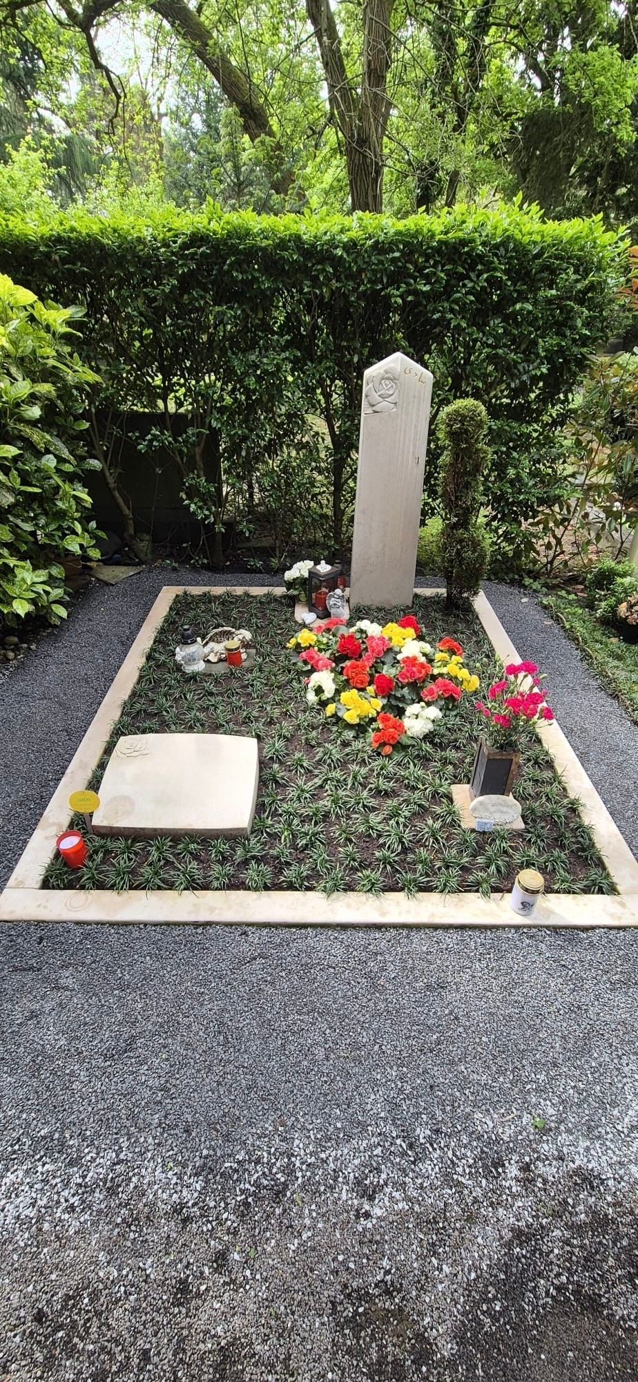 Kleiner Grabstein mit Blumen auf einem Friedhof, umgeben von Kies und Hecken.