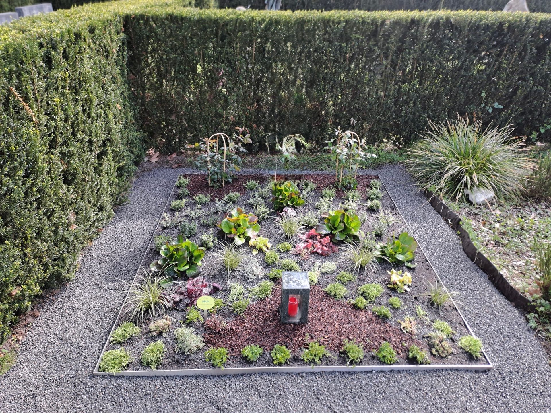 Grab mit bunten Blumen und Grünpflanzen, eingefasst von Kies und Hecken.