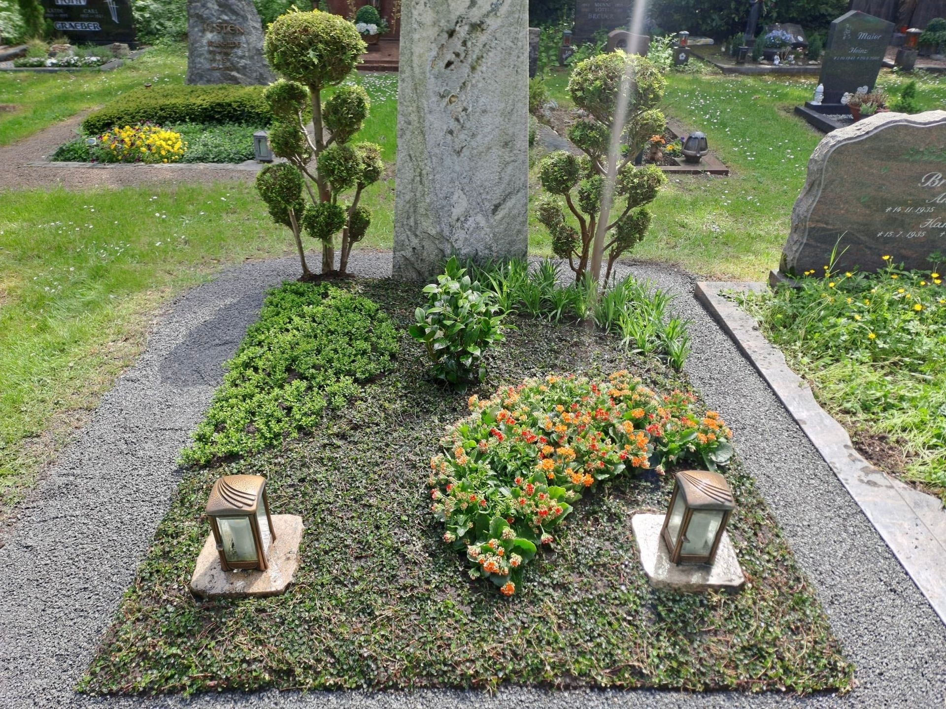 Kleines, landschaftlich gestaltetes Grab mit grüner Bodenbedeckung, Blumen und zwei Laternen neben einem Grabstein.