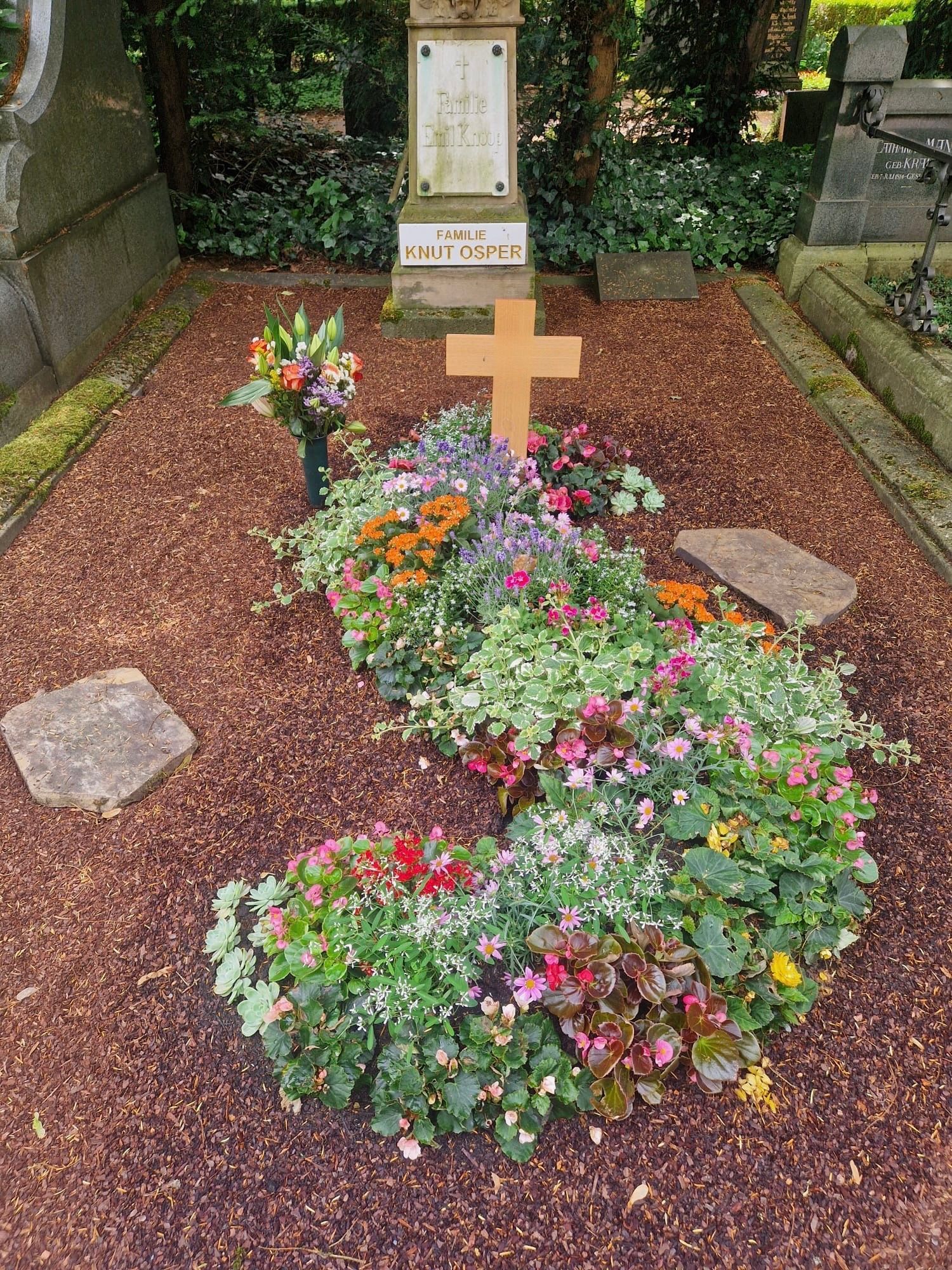 Ein mit Blumen bedecktes Grab mit einem Holzkreuz und einer Gedenktafel in einem Friedhofsgarten