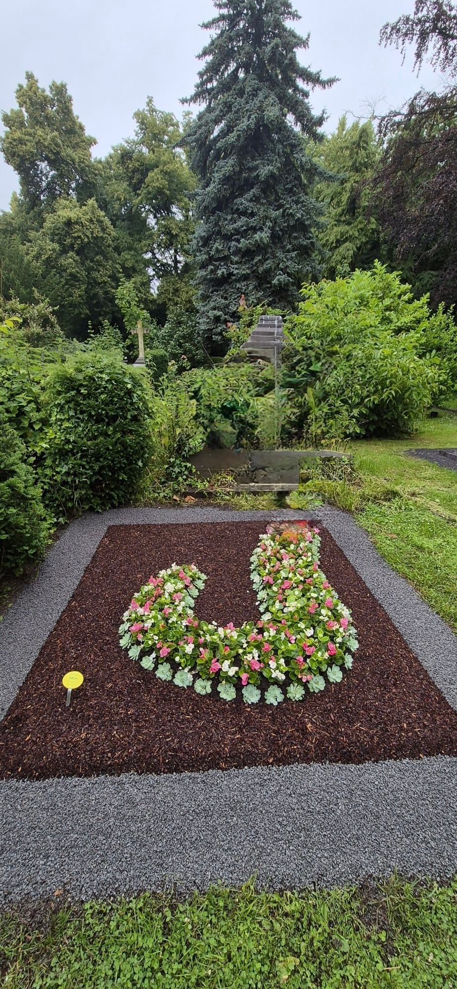 Blumenbeet als Gedenkstätte auf einem Friedhof, mit farbenfrohen, herzförmigen Blumen in einem dunklen Kiesrand.