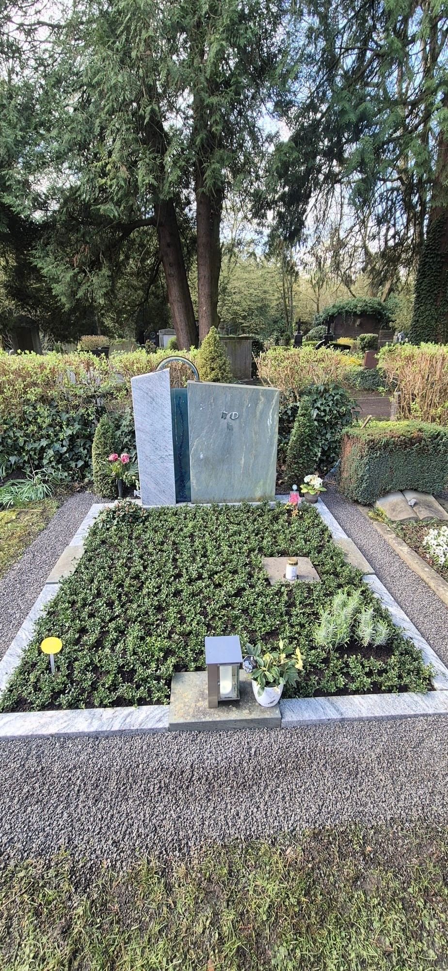 Grabstätte mit einem polierten grauen Grabstein auf einem Friedhof, umgeben von Kies, Blumen und Bäumen.